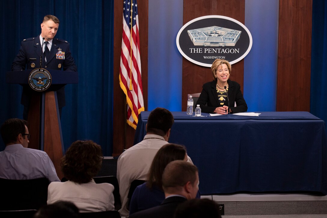 A woman sits behind a desk on a podium and speaks to reporters as a man in a military uniform stands next to her.