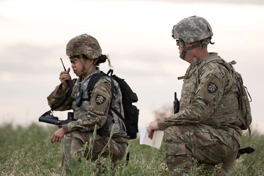 Two soldiers stand in a field, one communicating by radio.