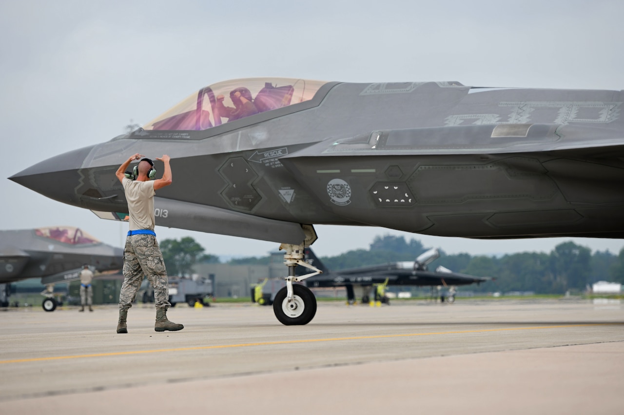 A pilot in a cockpit makes hand signals to an airman on the ground.