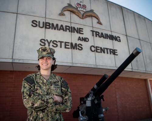 A female Sailor smiles while posing for a photo in front of a brick building with the words "Submarine Training and Systems Centre" written on the building.