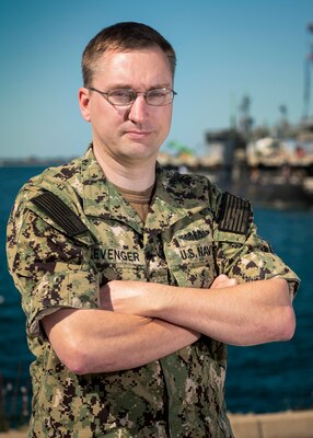 A male Navy Sailor poses with his arms folded, outside in front of a Navy pier in uniform.