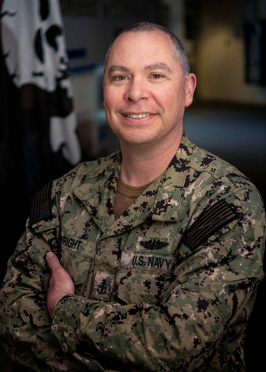 A male Navy Sailor stands with his arms folded, smiling for a photo in uniform.