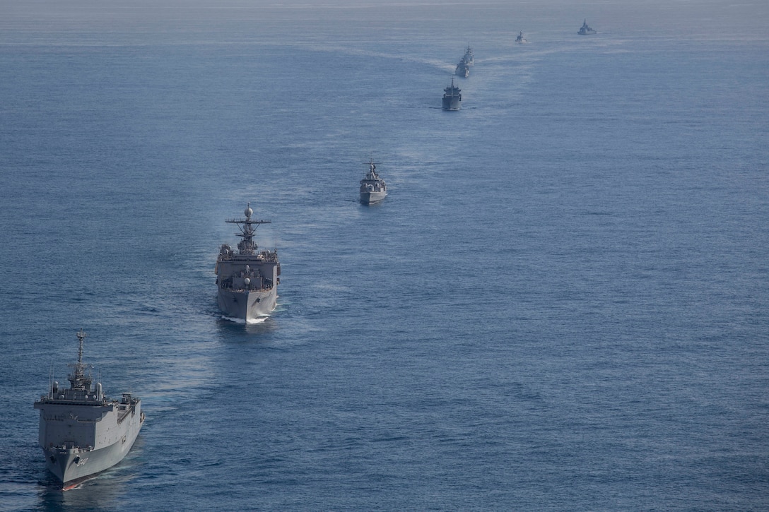 Military ships sail in a line through the ocean.