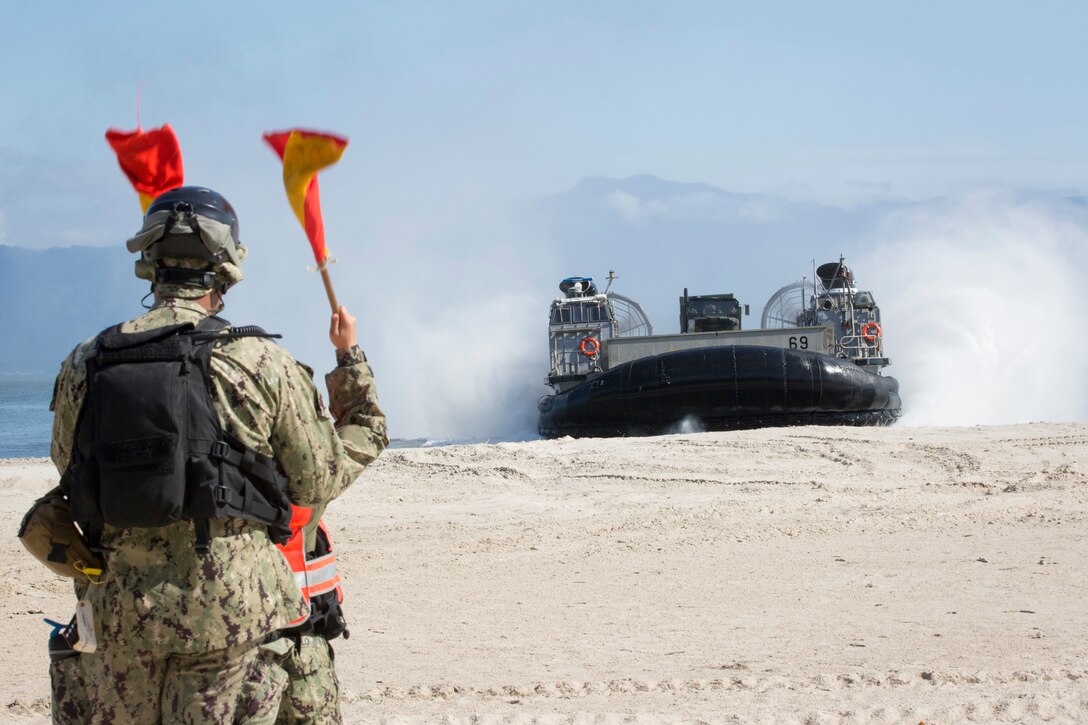 Sailors guide a military landing craft onto a beach from the ocean.