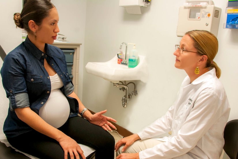 A pregnant woman sits on an exam table and speaks to a female health care provider.