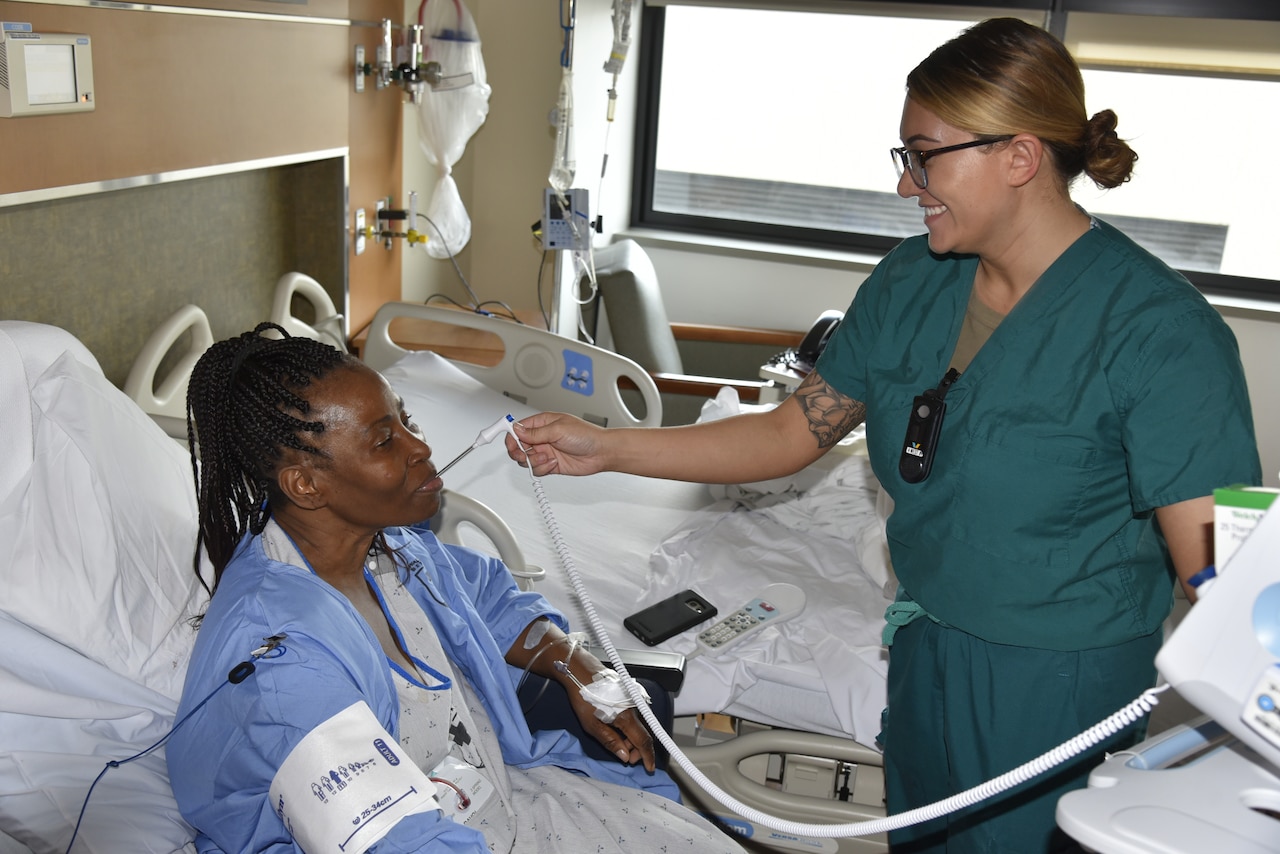 A nurse in blue scrubs holds a thermometer in the mouth of a patient who is in a hospital bed.