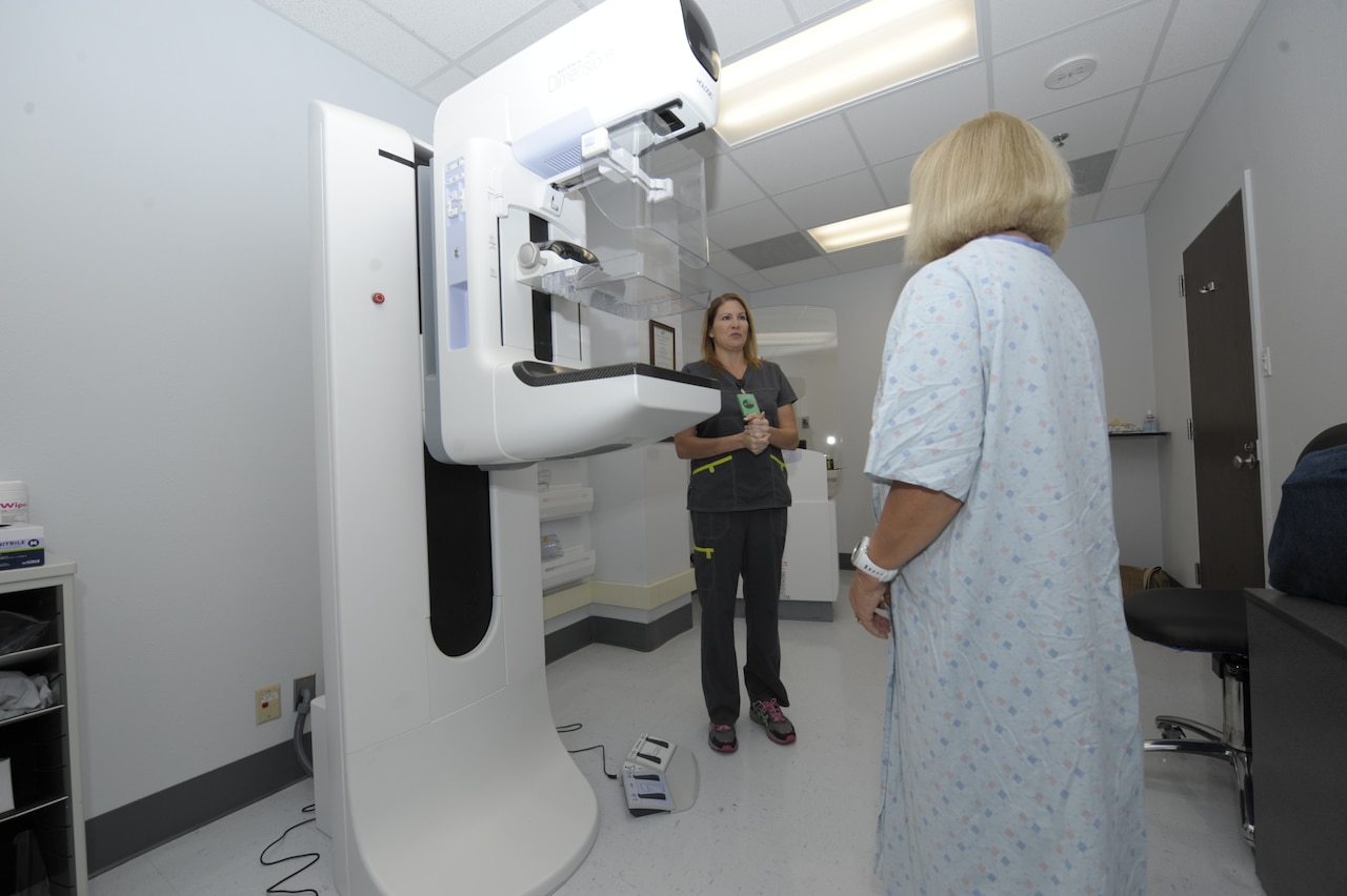 A female patient in a hospital gown talks with female health care provider wearing gray scrubs. A diagnostic machine is between them.