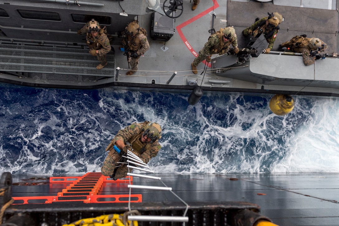 A service member climbs a ladder to get on board a landing platform while others watch from below.