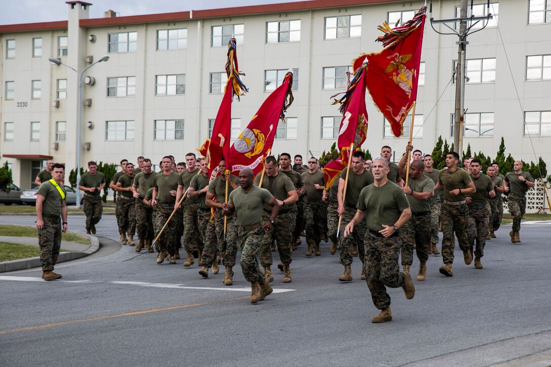 A large group of Marines run down a street; some holding flags.