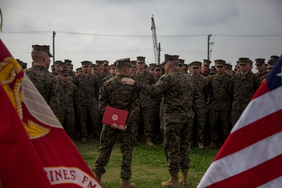 Two Marines stand with the backs facing the camera in front of a large group of Marines.