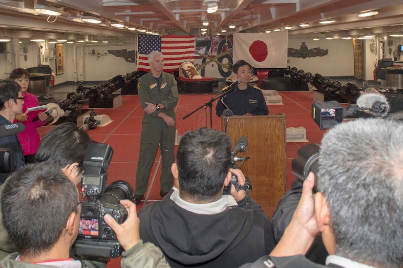 Two men in military uniform stand near a lectern. Behind them are the U.S. flag and the Japanese flag.