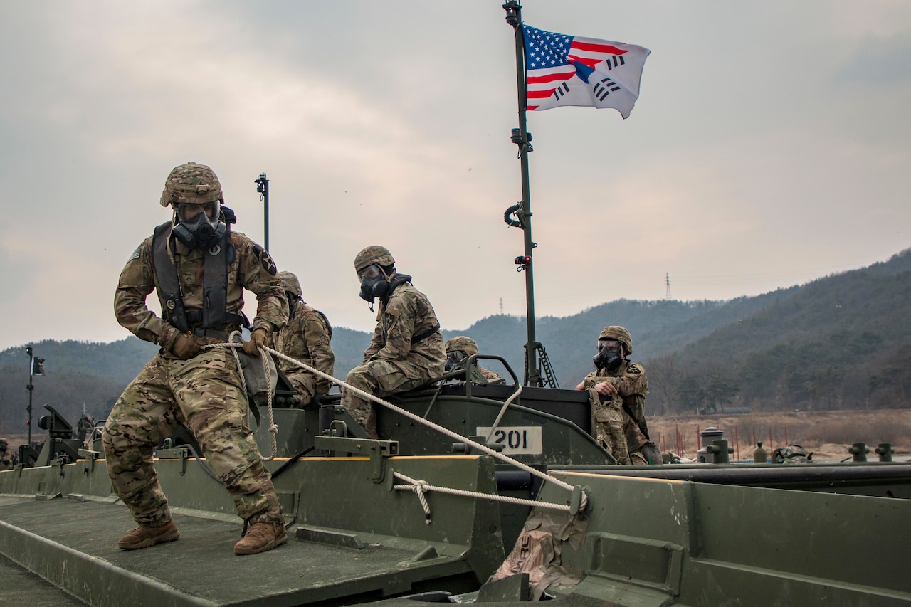 Military personnel wearing gas masks perform operations on waterborne craft.