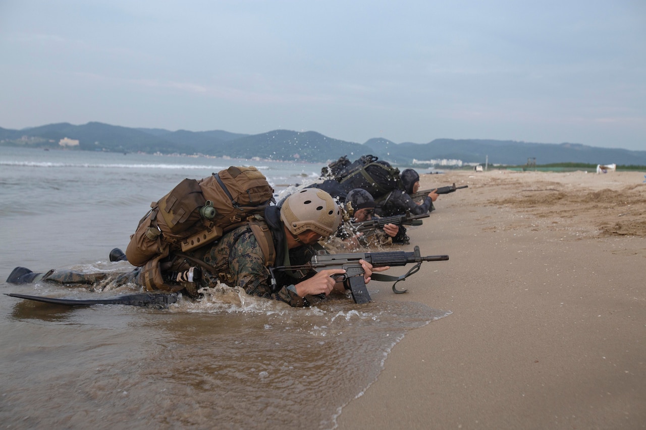 Military personnel with rifles crawl onto a beach from the water.