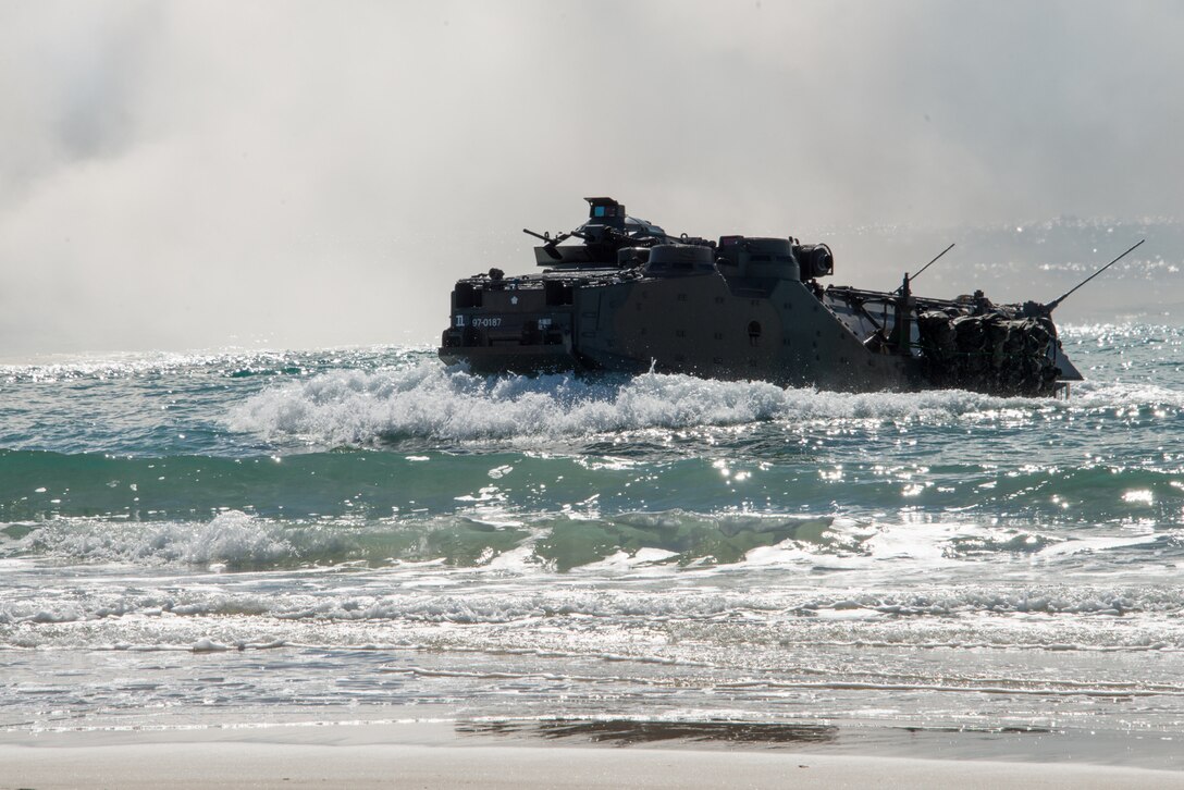 A large military vehicle plows through the water and approaches a beach.