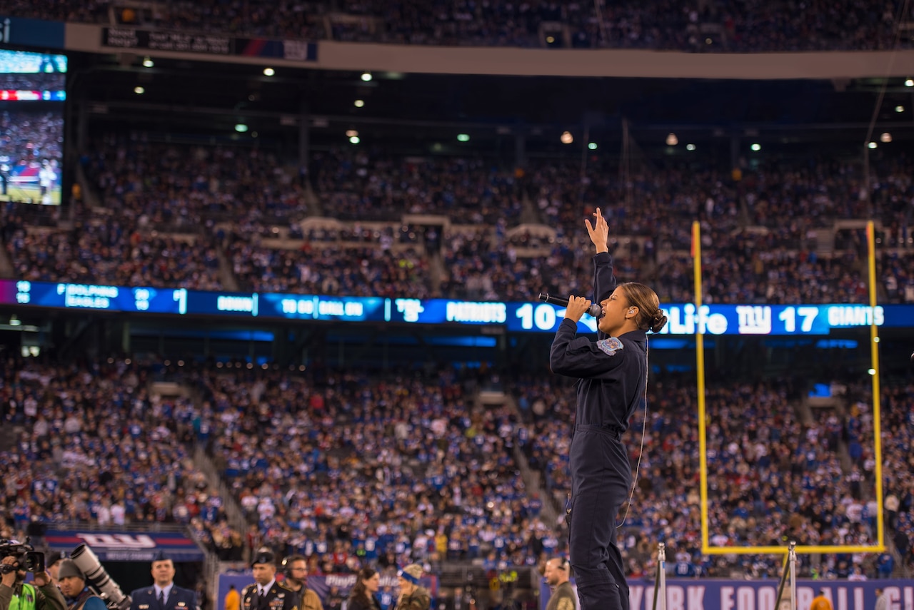 A singer in an Air Force flight suit standing in front of a crowded stadium holds one hand in the air and another to her mouth with a microphone.