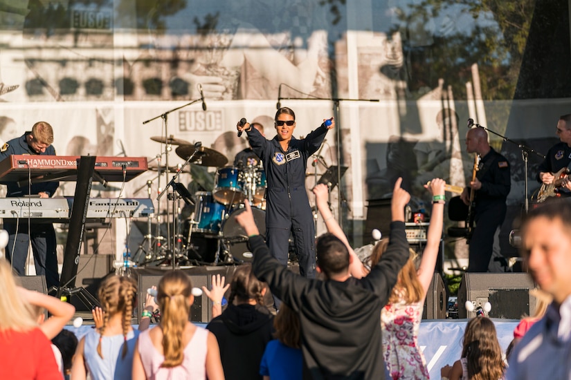 A singer on stage in a flight suit points both fingers toward the crowd looking at her.