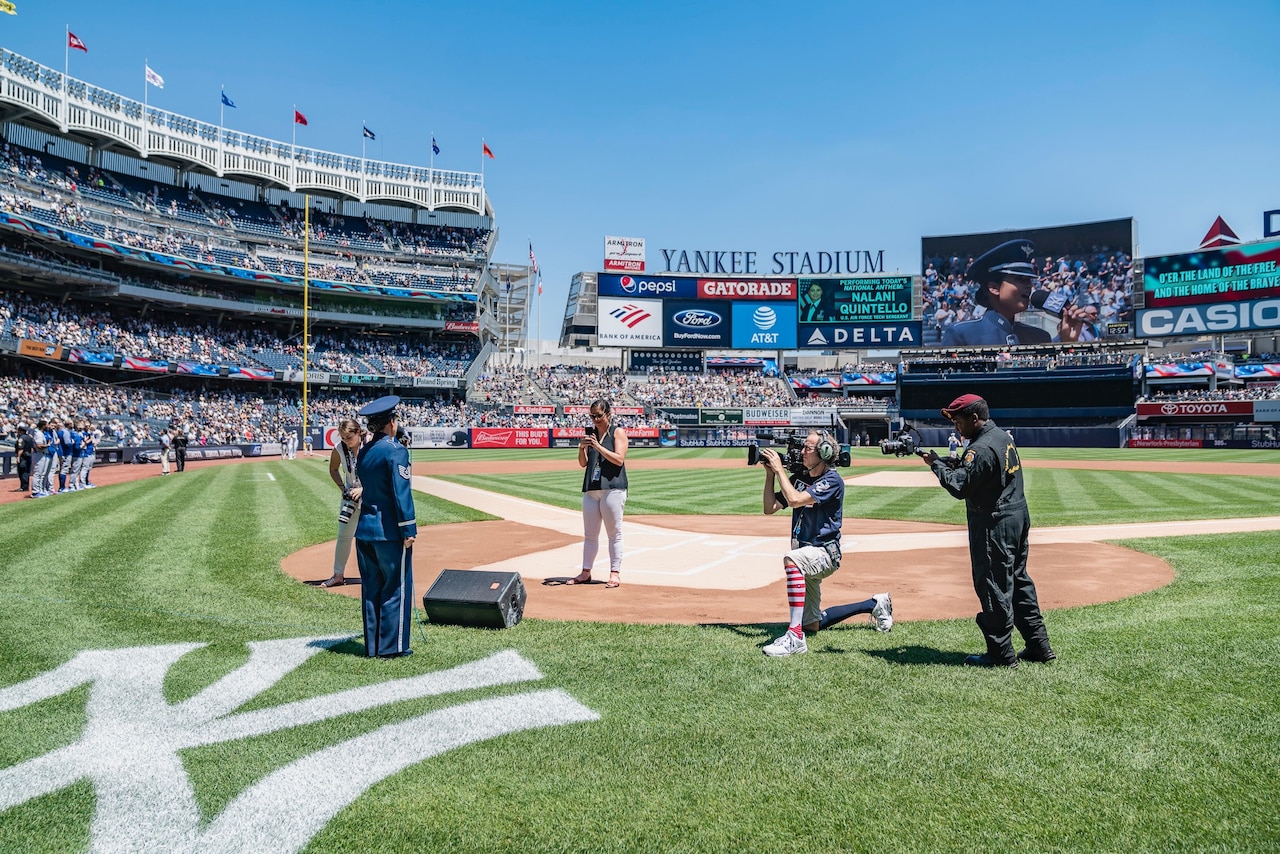 An airman in dress blues prepares to sing near the New York Yankees logo on the field at Yankees Stadium while four cameramen film her.