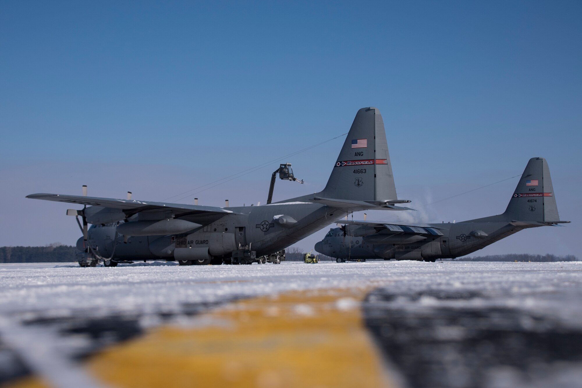 A photo of two C-130 aircraft and a deicing truck on the flight line in early morning.