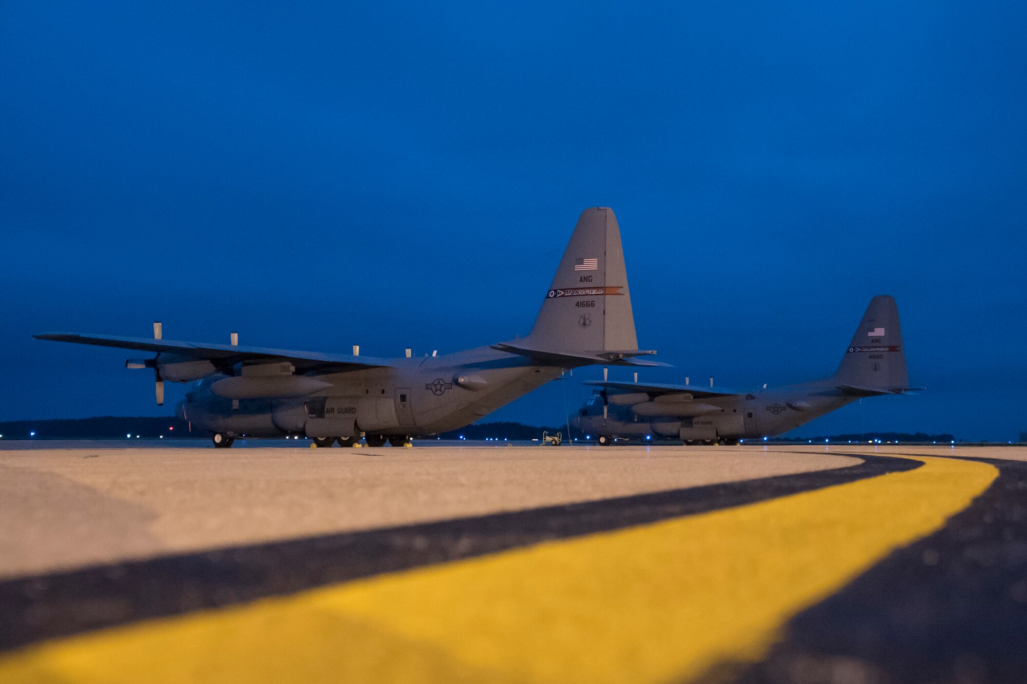 A photo of two C-130's on the flight line early morning against a dark blue sky.