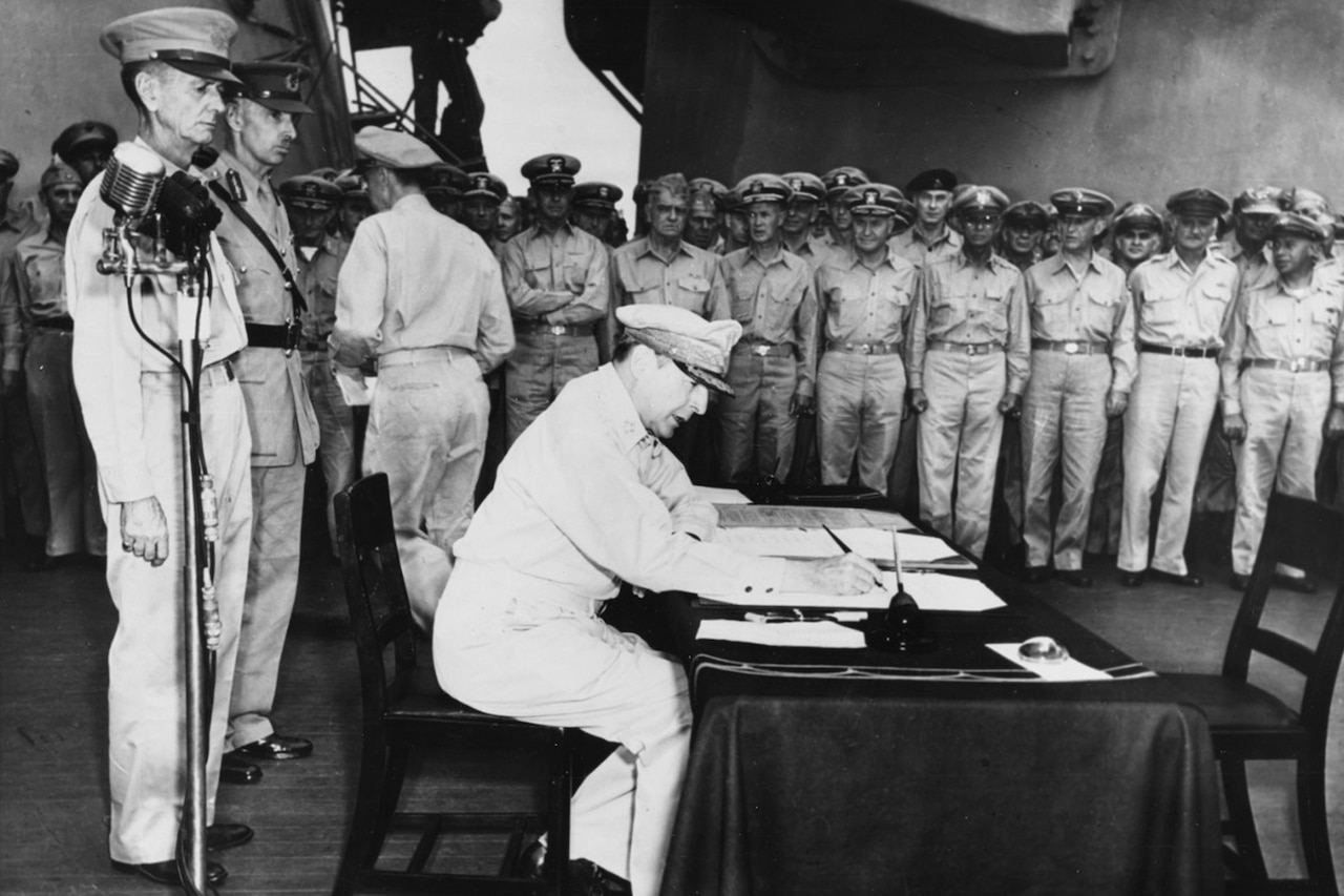 A soldier sitting in a chair at a table, signs the Japanese surrender document that ended World War II. Two other service members in dress uniform stand behind him near a microphone, and others stand off to the side.