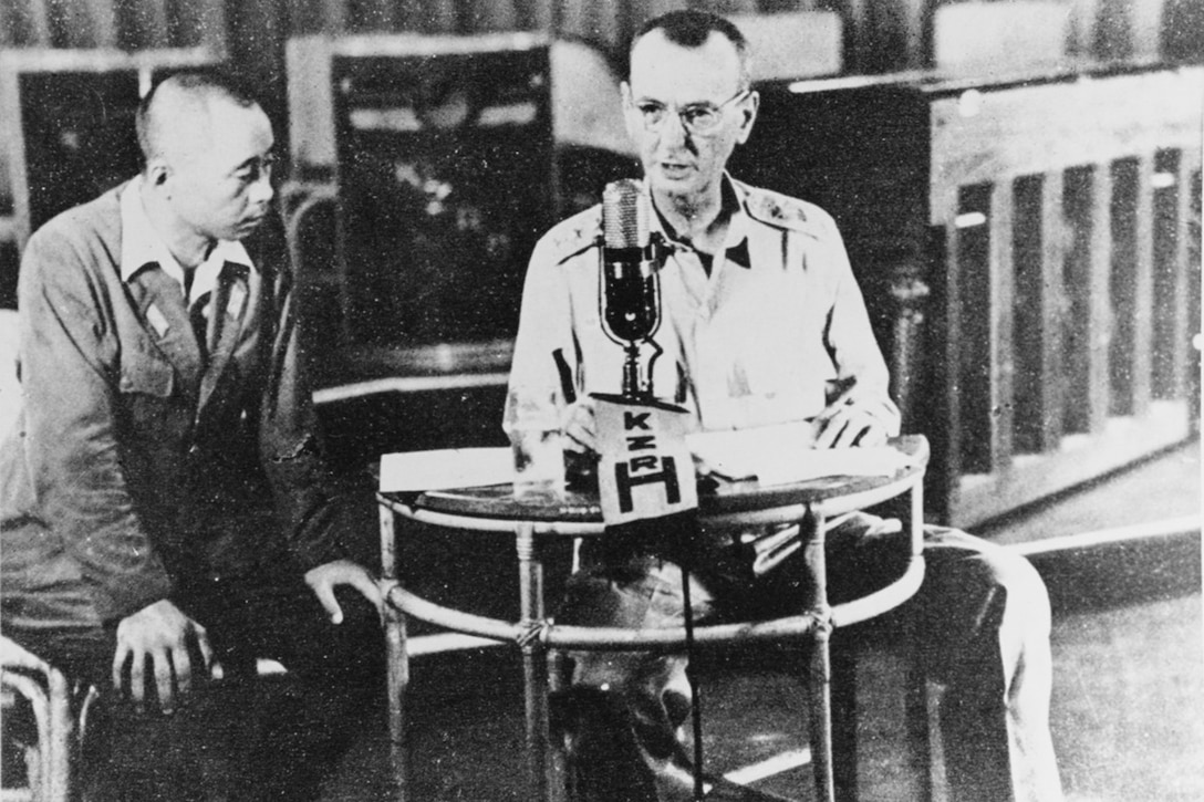 Army Lt. Gen. Jonathan Wainwright,  thin and wearing a button-down shirt and khaki pants, sits at a small table in front of a microphone as he broadcasts a message surrendering to the Japanese. A Japanese man sits beside him, looking at papers on the table.