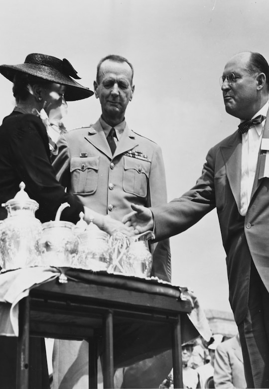 A woman wearing a hat shakes a man’s hand behind a table with several tea kettles on it. A second man in uniform stands between them.