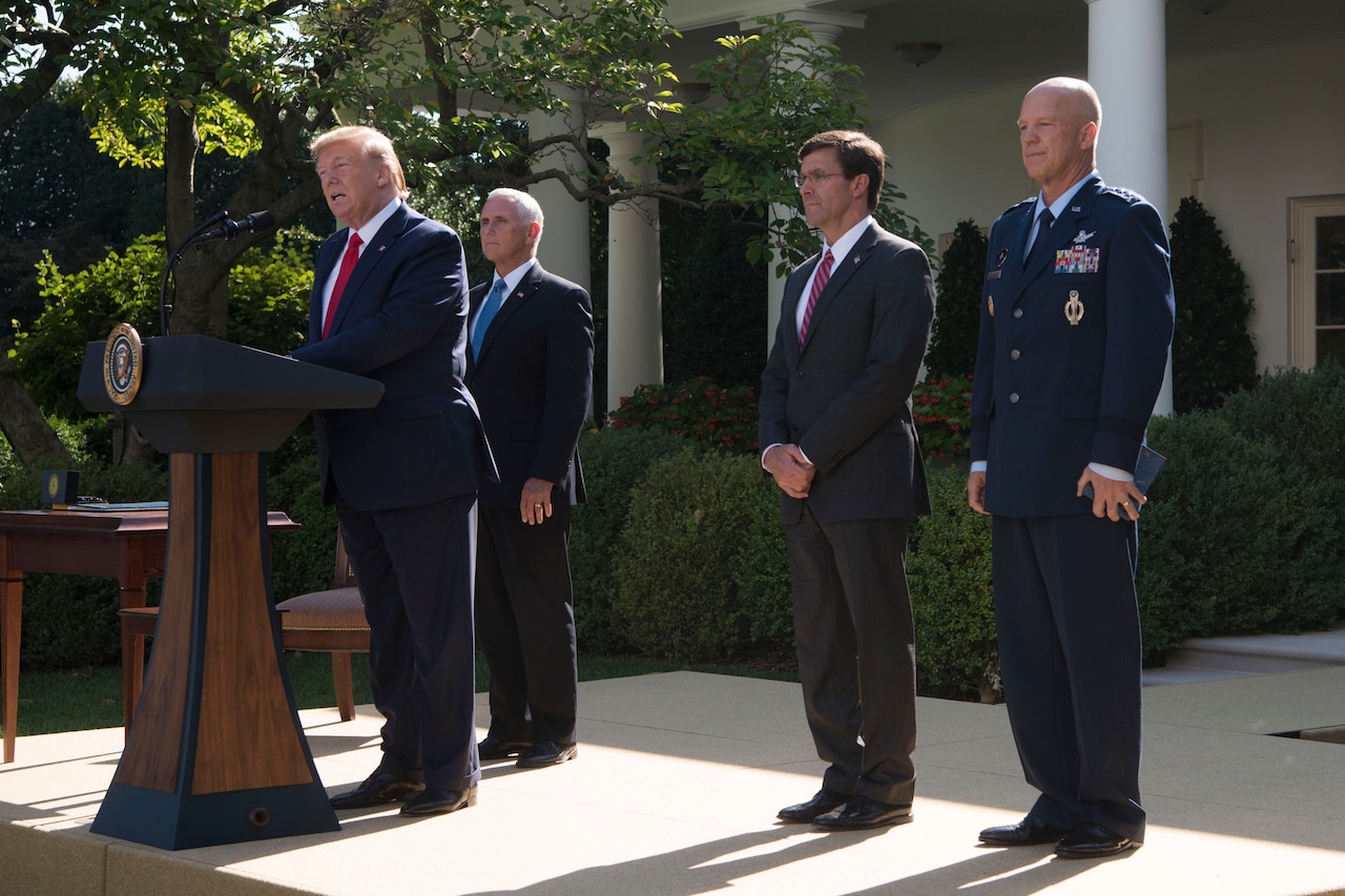 President Donald J. Trump speaks at a podium with two men in suits and an Air Force general standing behind him.