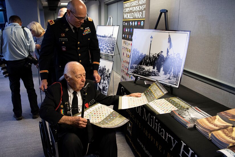 A man in a military uniform stands behind a man seated in a wheelchair.  The man in the wheelchair looks at documents related to World War II.