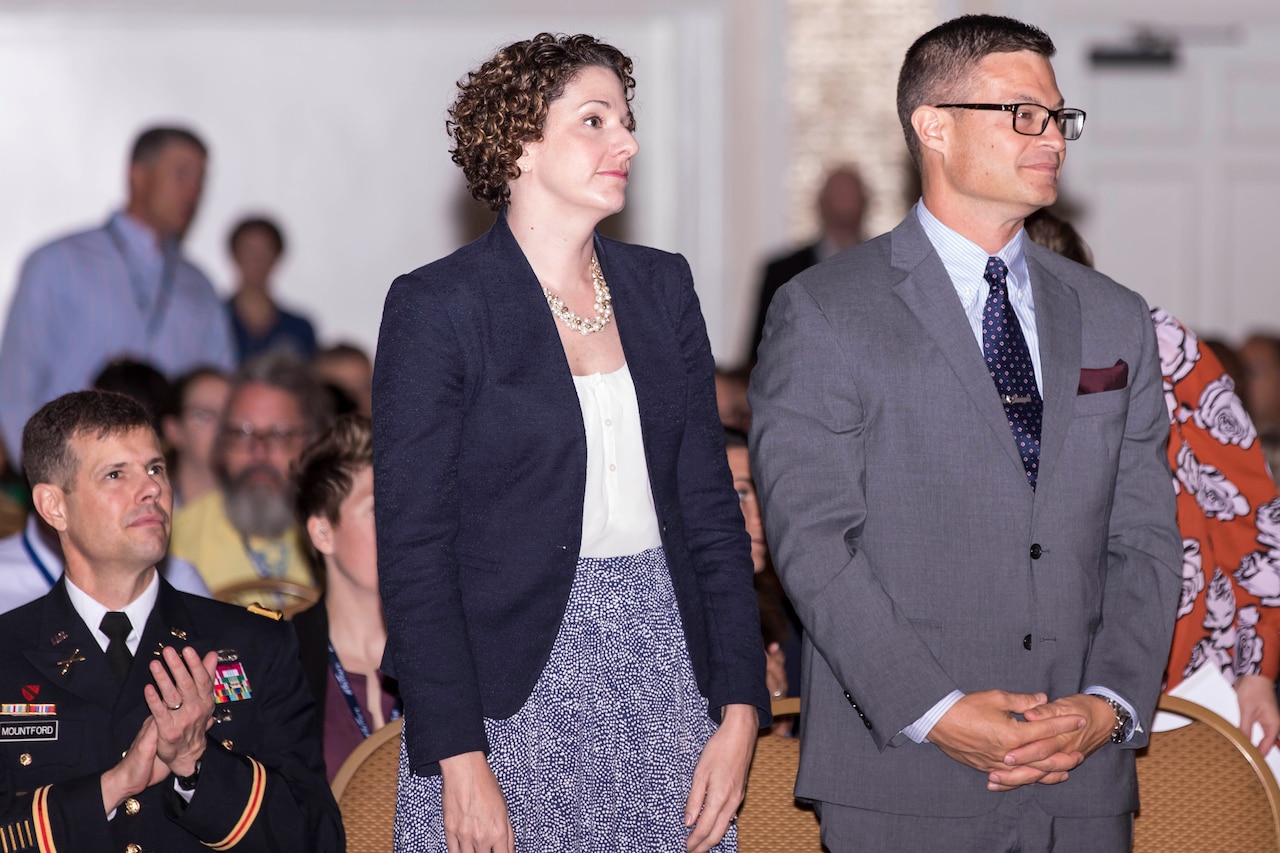 A woman and man stand while other sit around.