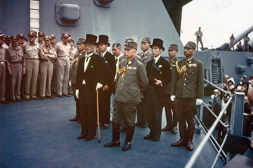 Japanese civilians in formal attire and officers in uniform stand on battleship deck with Allied service members standing nearby.