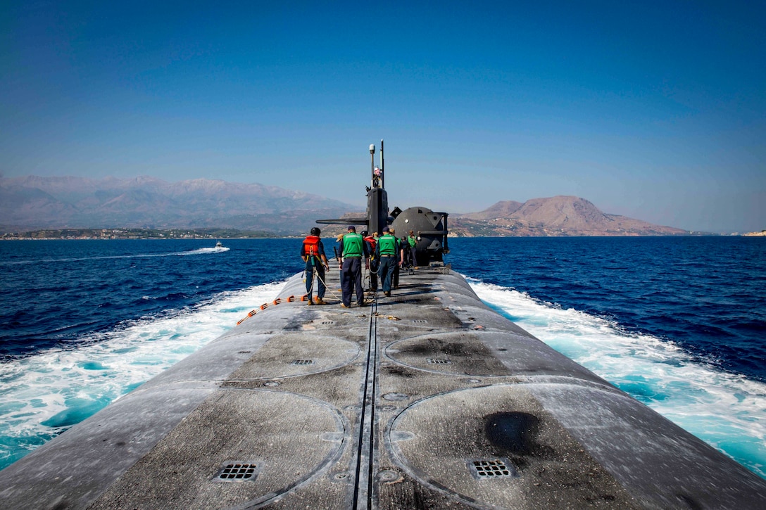 Sailors stand on top of a submarine as it travels through waters.