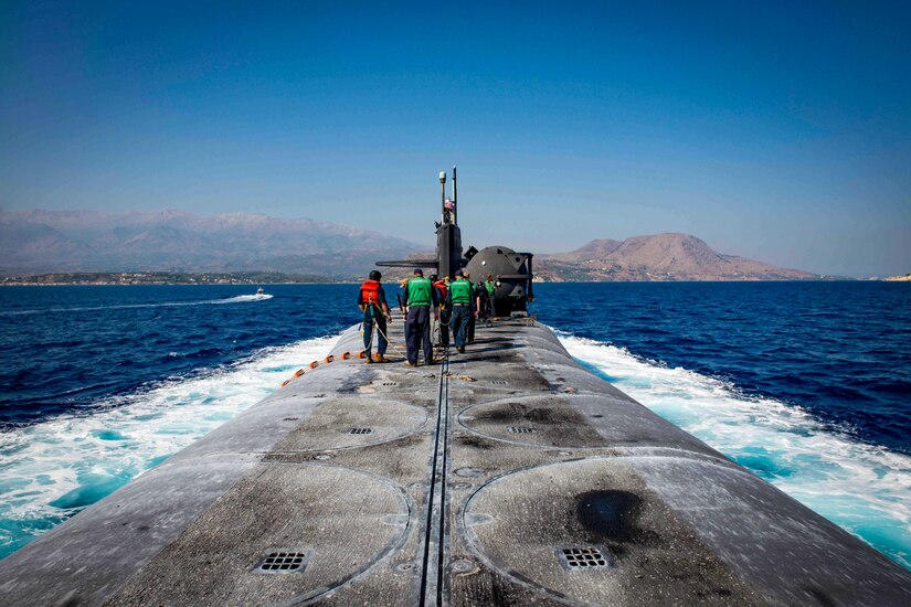 Sailors stand on top of a submarine as it travels through waters.