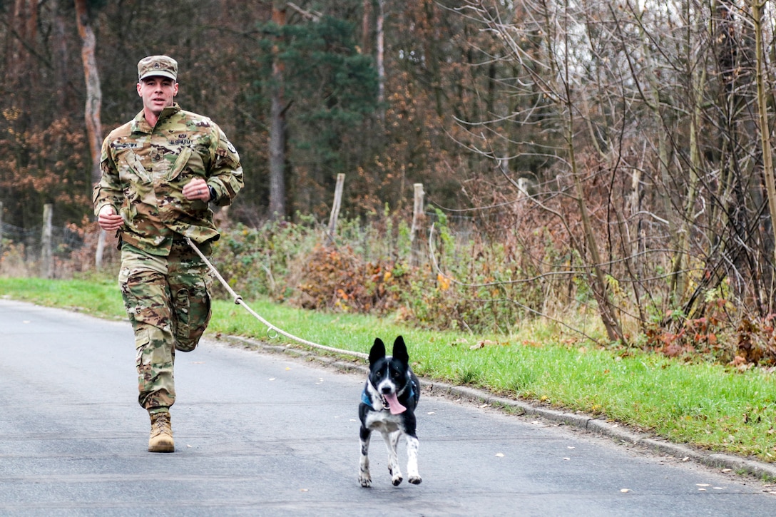 A soldier runs with a small black-and-white dog on a road.