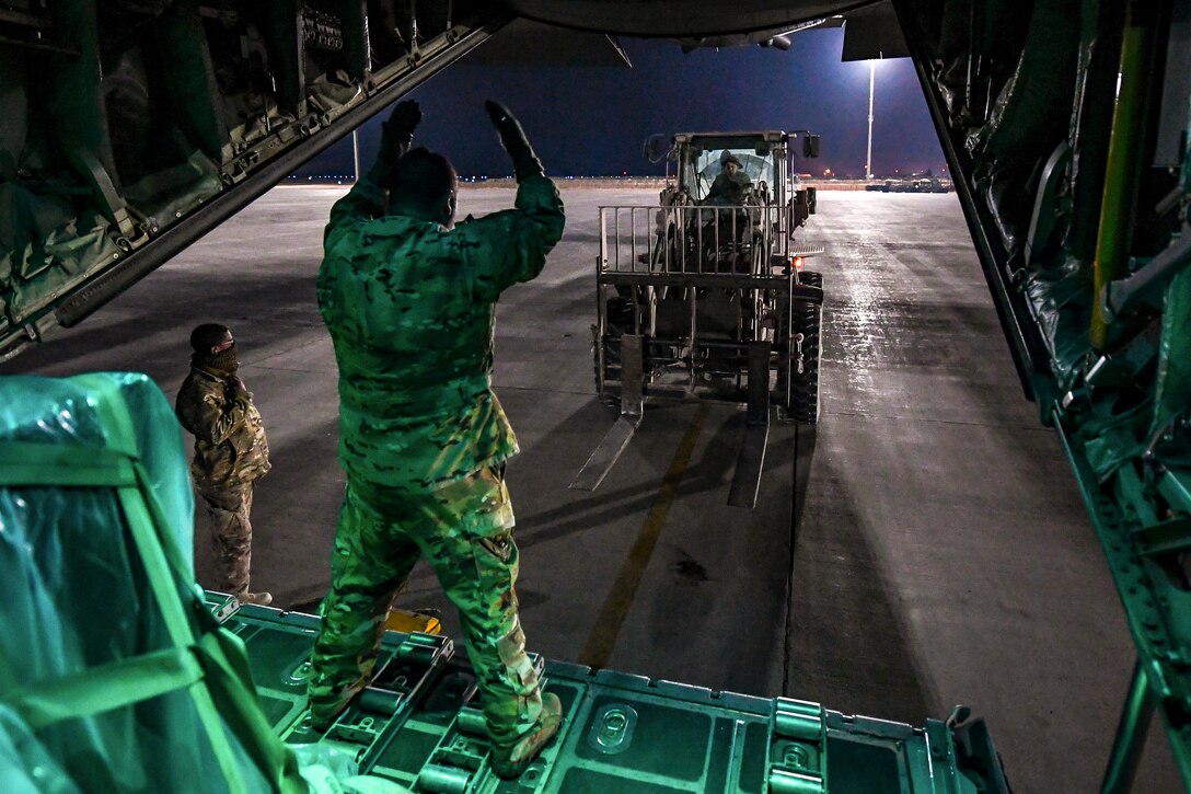 An airman in an open aircraft gestures to a forklift driver on a flightline.
