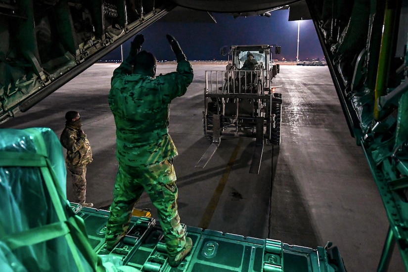 An airman in an open aircraft gestures to a forklift driver on a flightline.