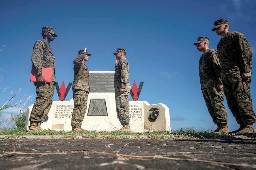 Two Marines stand and face each other with right hands raised by a memorial atop a mountain as three Marines stand by.