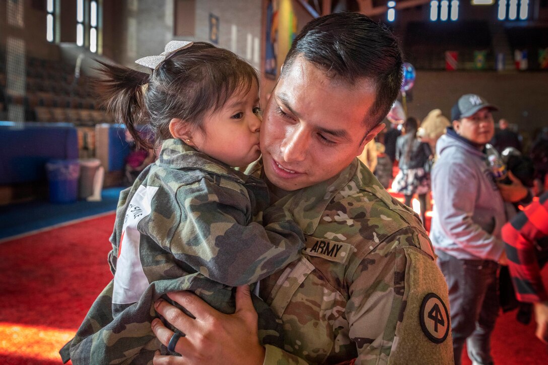 A soldier is kissed by his young daughter as he holds her in his arms.