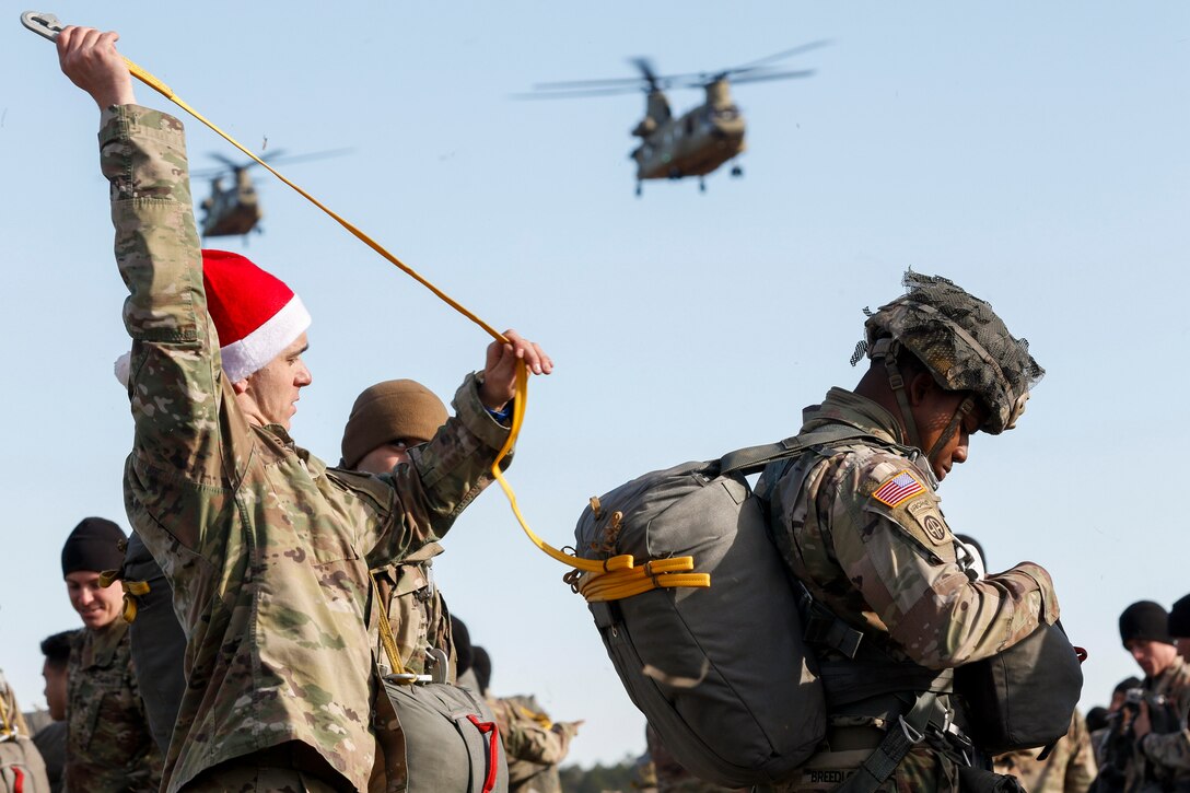 A soldier in a Santa hat inspects the parachute of a solider standing in front of him in a field as helicopters hover above.