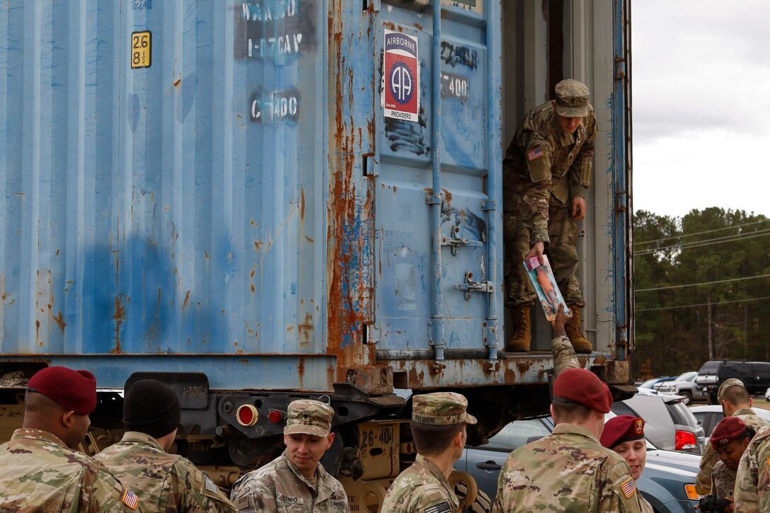 A soldier standing on the ground outside passes a toy to a soldier standing in a shipping-type container.