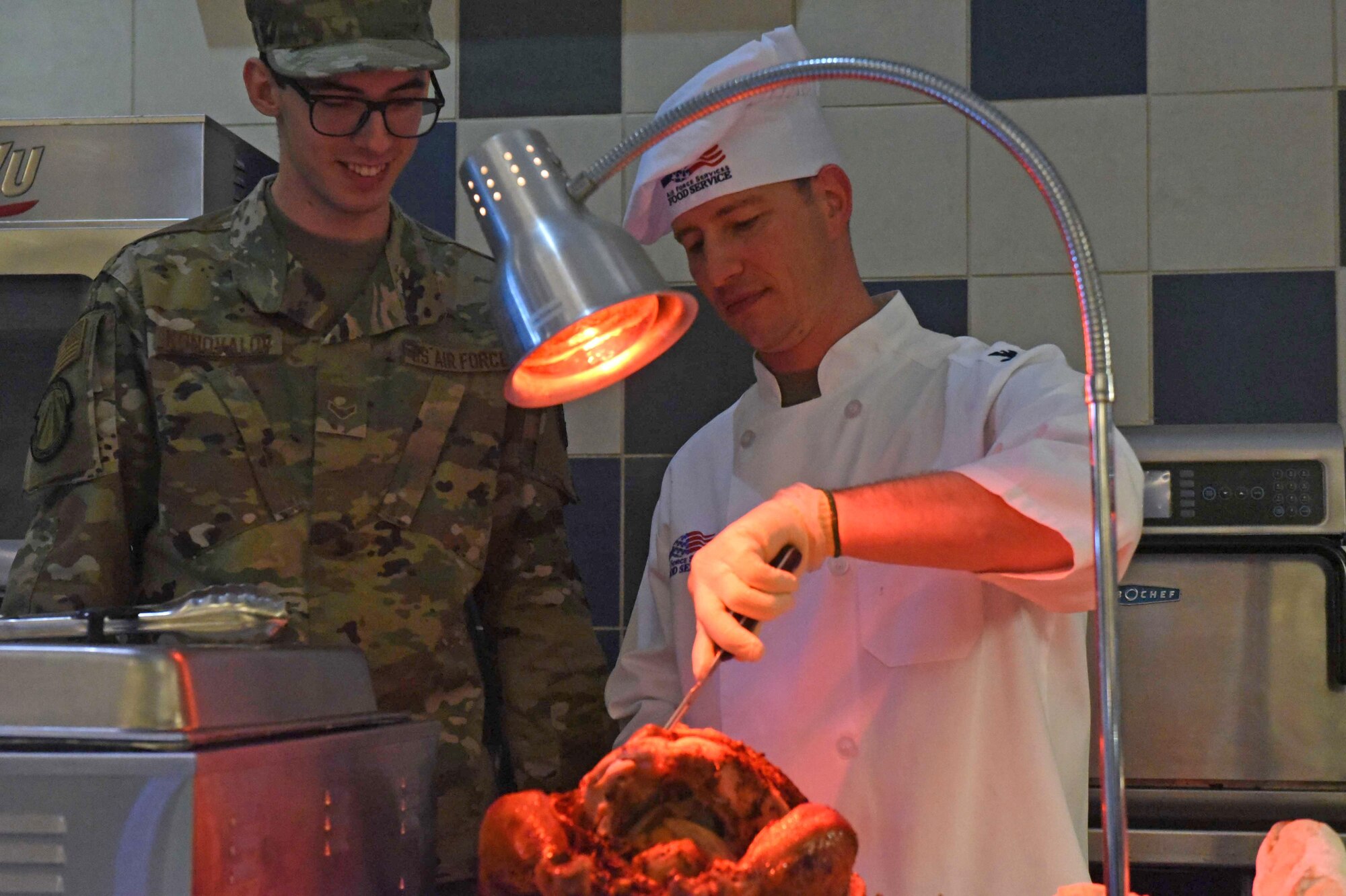 Airmen in chefs coat serve meals in cafeteria line