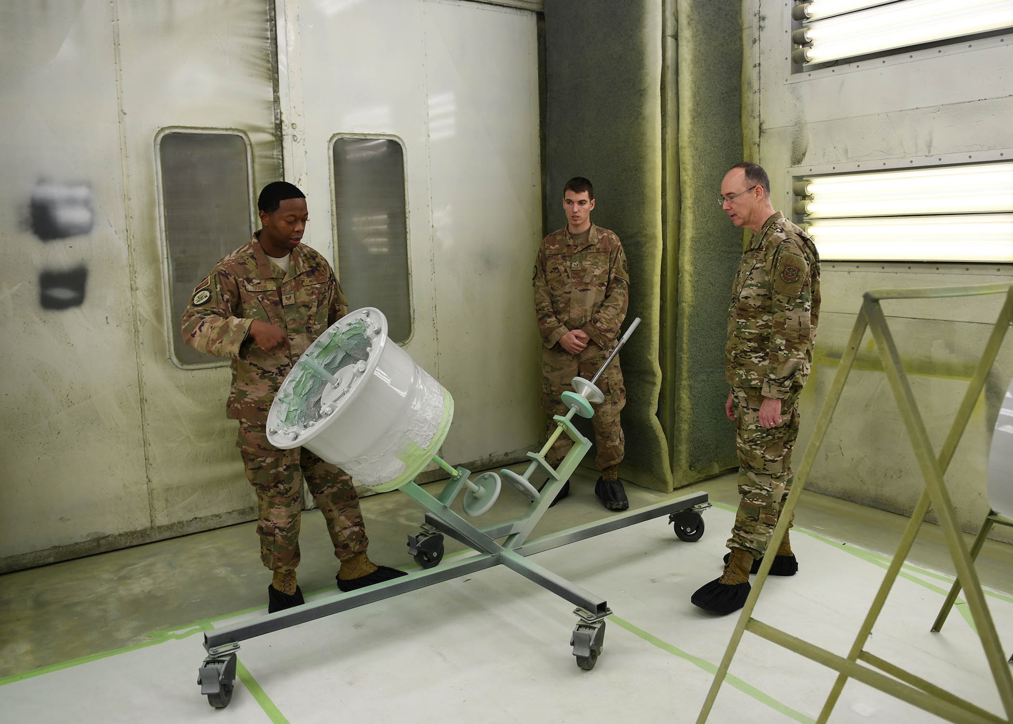 Three Airmen look at a piece of equipment in a room used for painting plane parts.