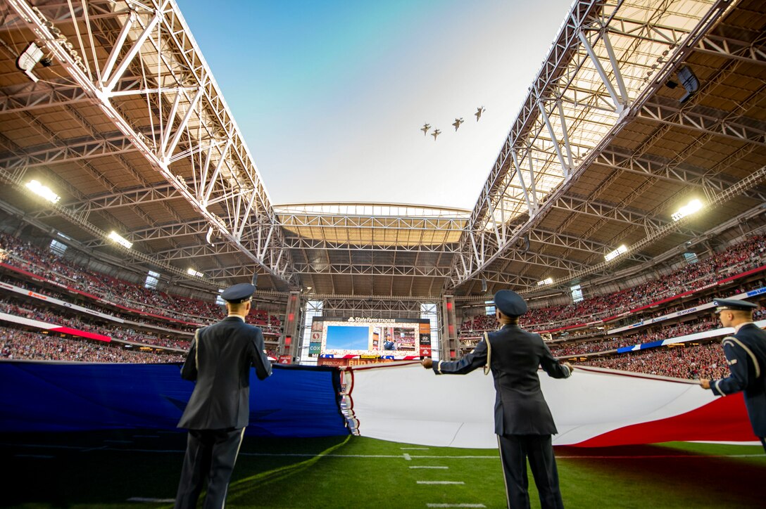 Airmen hold a giant American flag on a football field as four aircraft fly over the stadium.