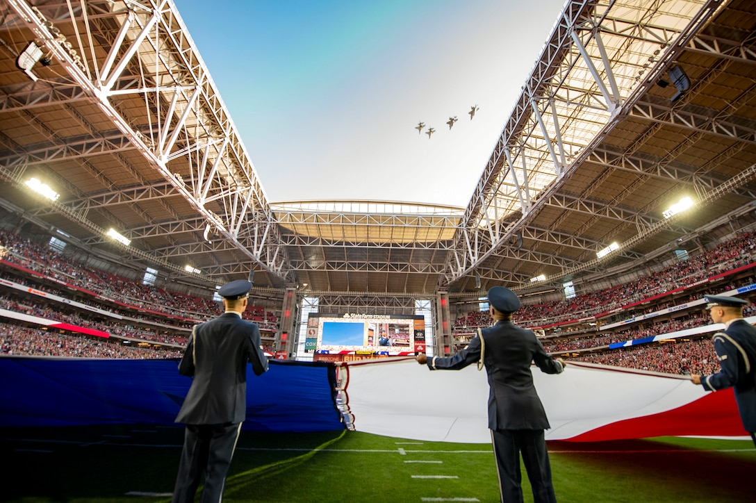 Airmen hold a giant American flag on a football field as four aircraft fly over the stadium.