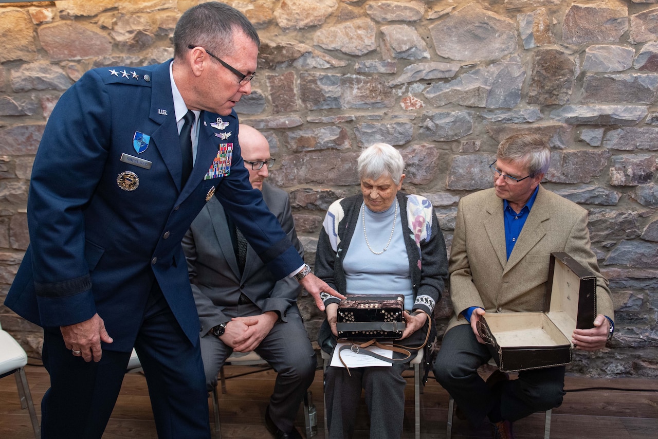 General points to accordion as two men and a woman inspect it.