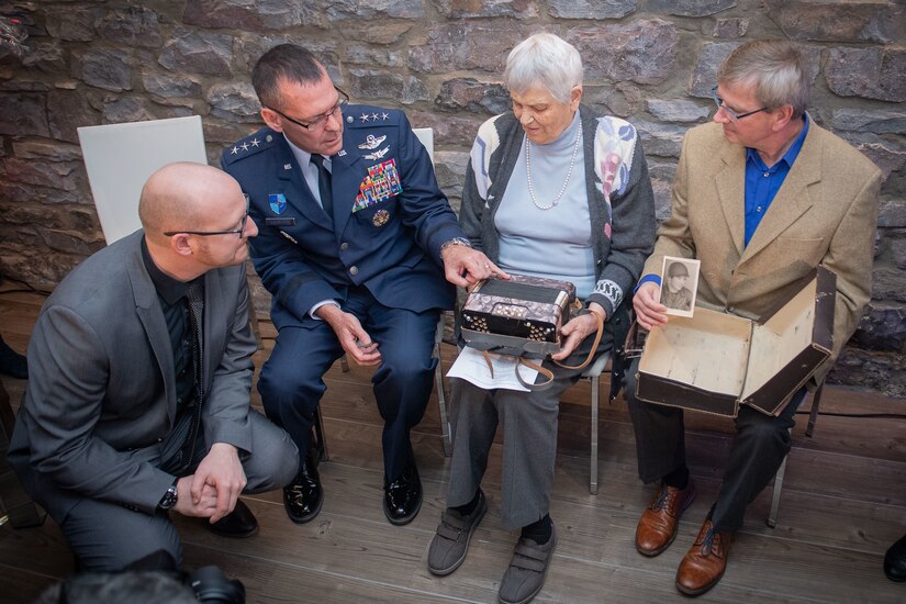 Three men and a woman sit in chairs as they look at an accordion found during World War II. One of the men holds a photo of a soldier above an album that is in his lap.