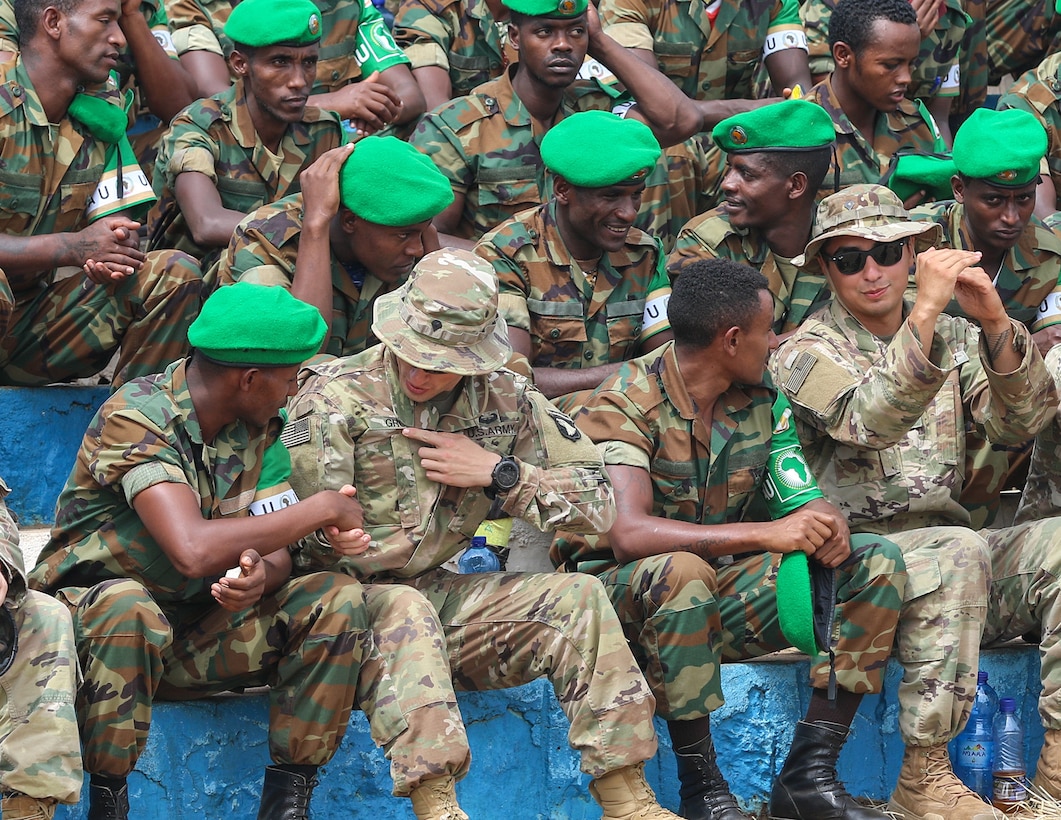 Soldiers in camouflage uniforms, some wearing bright green berets, talk while sitting in bleachers.