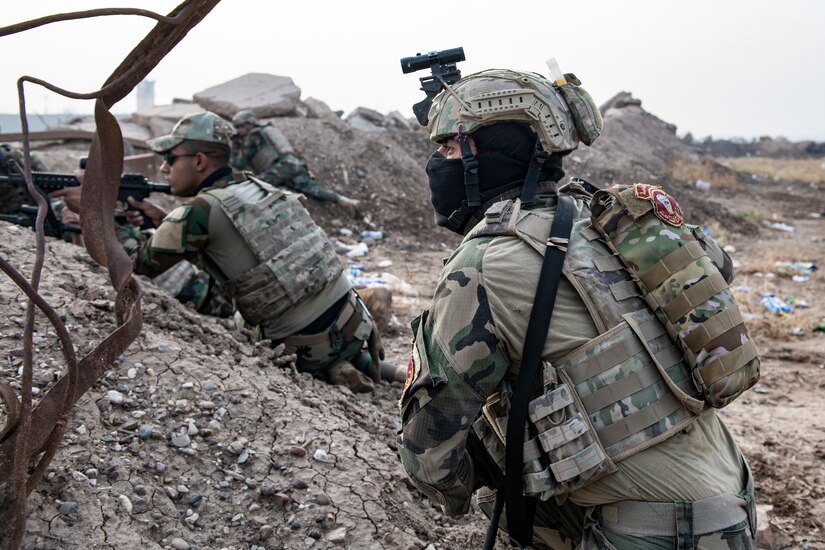 Soldiers lay protected behind a berm. One points a rifle.  Another looks over the berm.