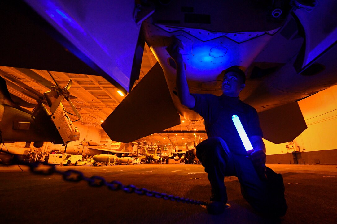 A sailor kneels under an aircraft while holding a blue light.