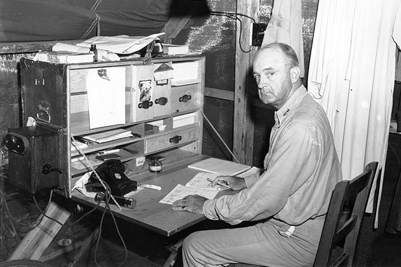 An older Marine looks over his shoulder at the camera as he sits at a work station in a tent.