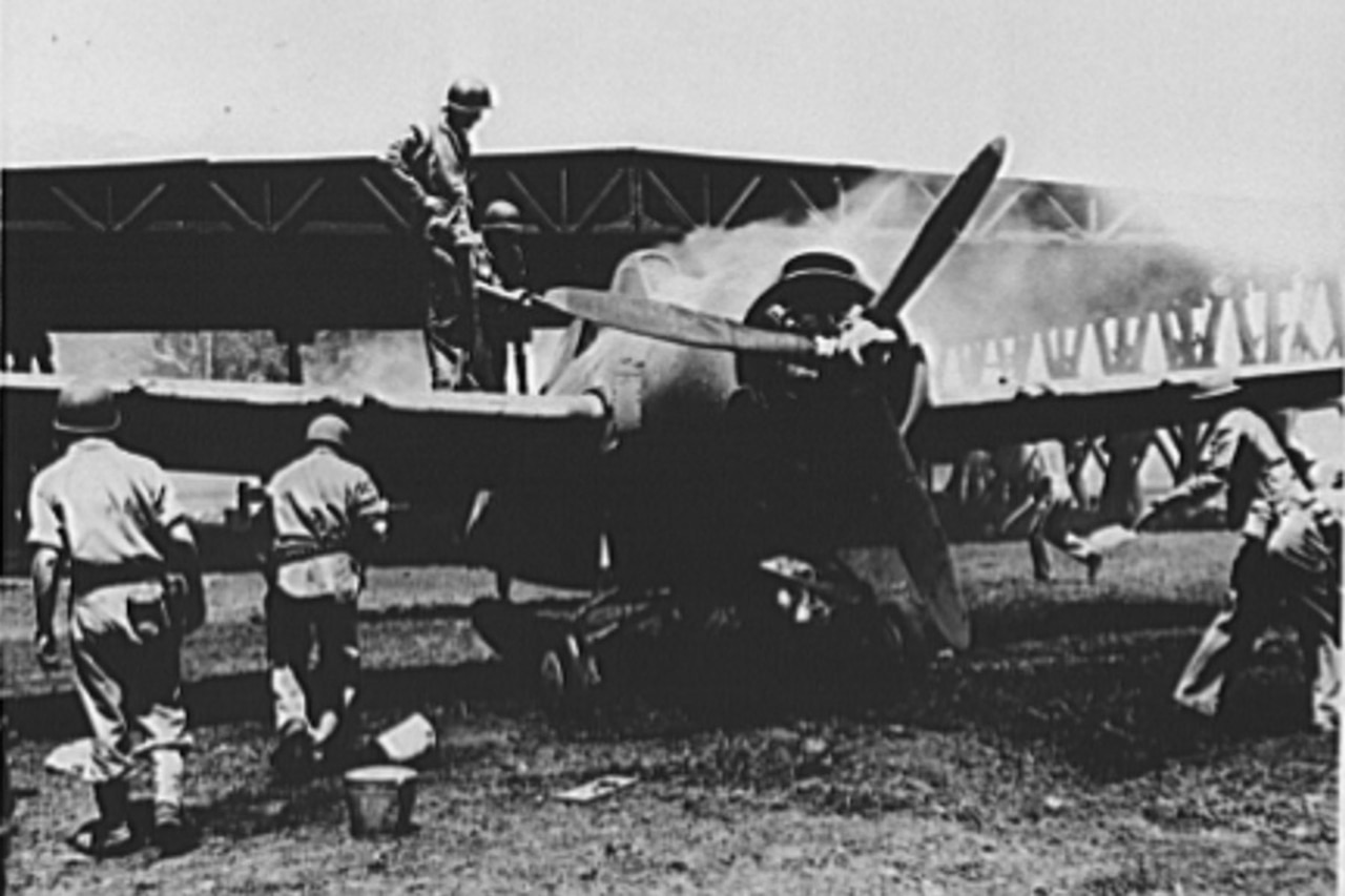 Marines on the ground put out a propeller airplane fire as two men stand on its wing to take a look. A hangar is in the background.